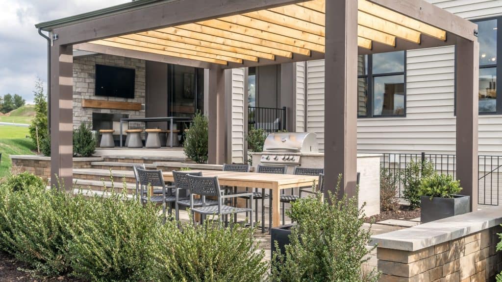 Modern outdoor patio with wooden pergola, dining area, built-in grill, and bar area with stools; surrounded by greenery and attached to a house with large windows—expertly designed by a top sunroom contractor Long Island, NY.