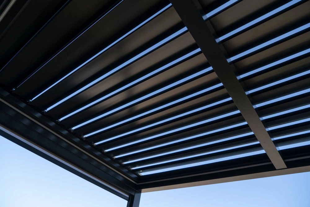 Close-up view from below of a modern black metal pergola with adjustable horizontal slats, partially open to let blue sky and sunlight through—perfect for Suffolk all season sunrooms or sunroom installation projects.