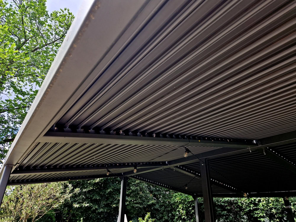 Close-up view from beneath a modern, corrugated metal patio roof supported by black metal beams—perfect inspiration for Sunroom Installation in Suffolk. String lights hang below, with lush green trees and foliage in the background.