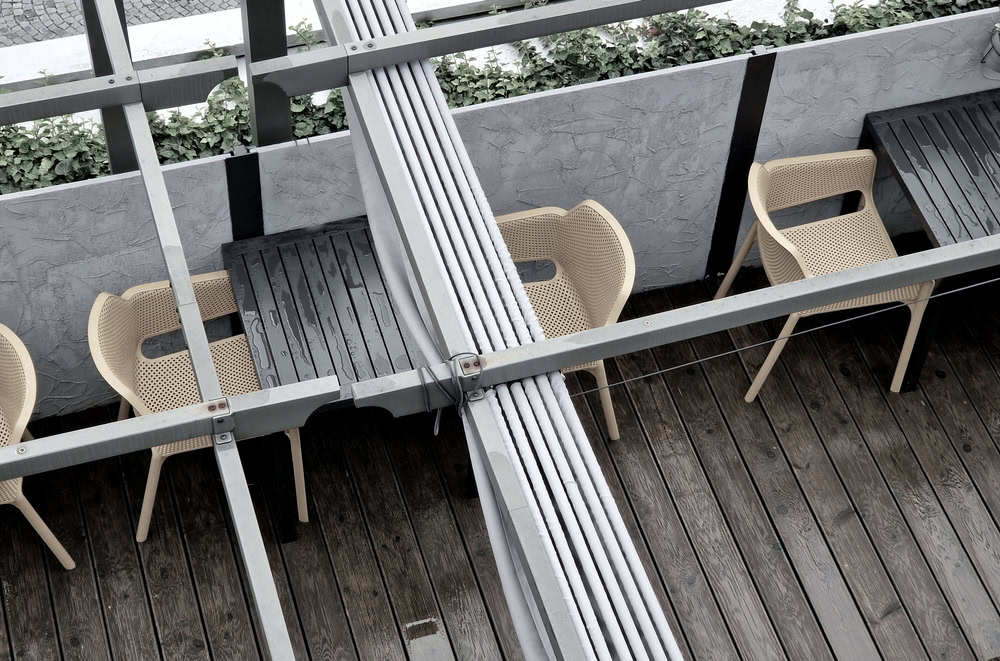 Outdoor seating area with beige woven chairs and black tables on wooden decking, viewed from above through a metal and wire frame structure. Green ivy lines a concrete wall in the background, evoking a relaxed Nassau vibe.