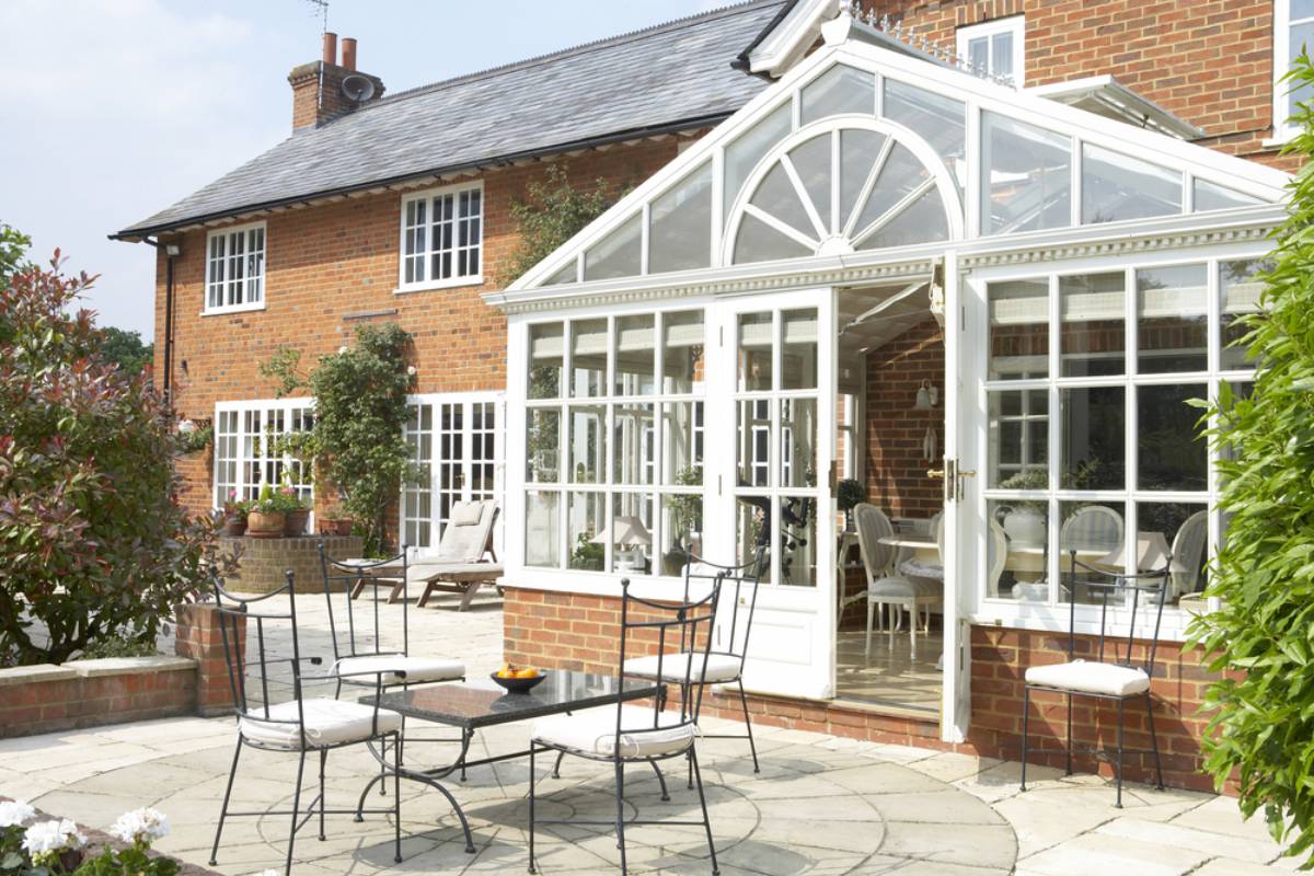 A sunlit patio with black wrought-iron furniture and white cushions sits outside a large Suffolk brick house, featuring a white-framed glass conservatory—perfect for those considering sunroom installation. Green plants and shrubs surround the area.