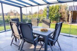 A modern glass sunroom by a top sunroom contractor Long Island, NY features a gray dining table with six cushioned chairs and a decorative centerpiece, offering relaxing views of the lush garden and trees outside.