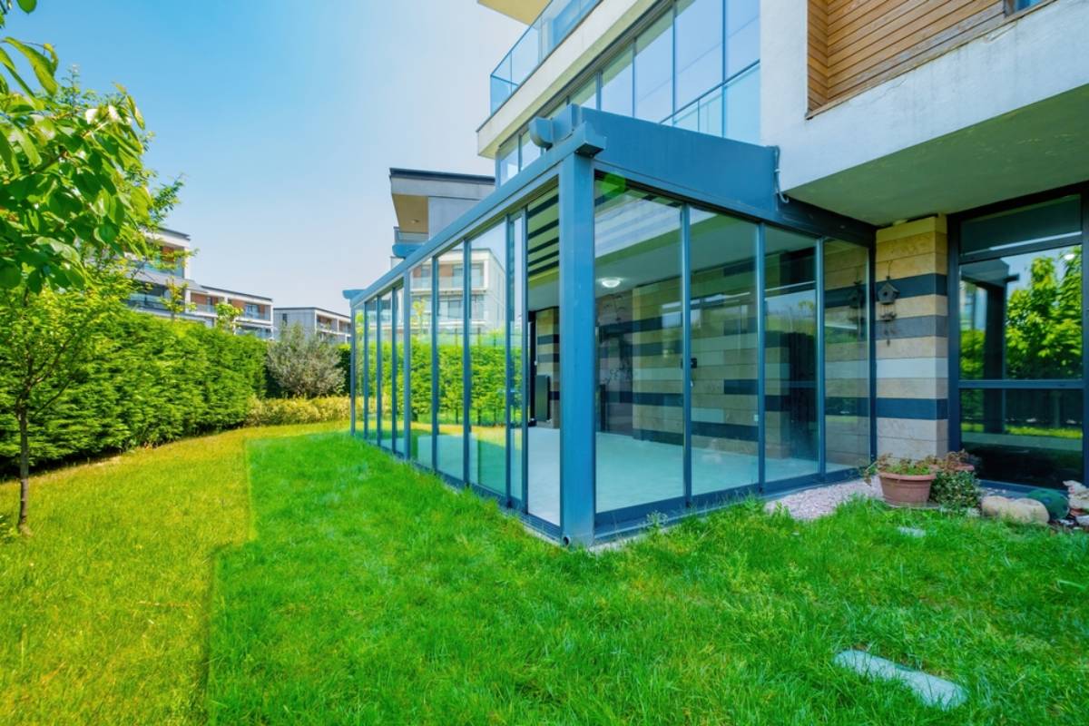 A modern glass-enclosed patio, designed for all season sunrooms, features a striped wall and overlooks a lush green lawn bordered by hedges and potted plants on a sunny day.