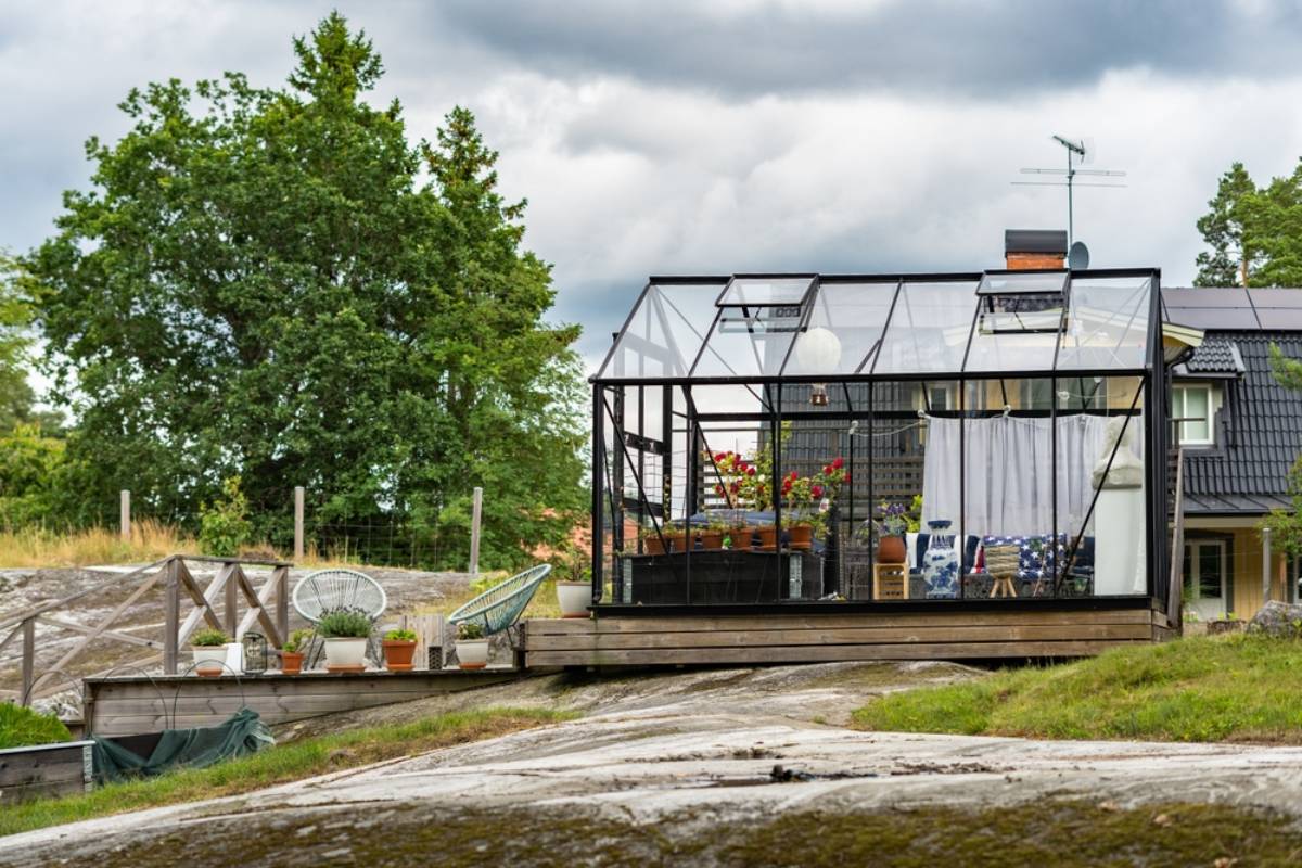 A modern greenhouse designed for all season sunrooms sits on a wooden deck attached to a dark-roofed Suffolk house. Potted plants and outdoor furniture decorate the deck, surrounded by trees and natural rocky landscape under a cloudy sky.