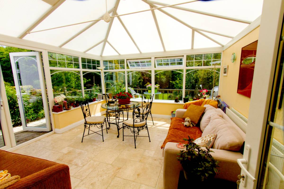 A bright conservatory, crafted with expert sunroom construction in Suffolk, features large glass windows, a round dining table, beige sofa, potted plants, a dog, and doors opening to a lush Long Island garden view.