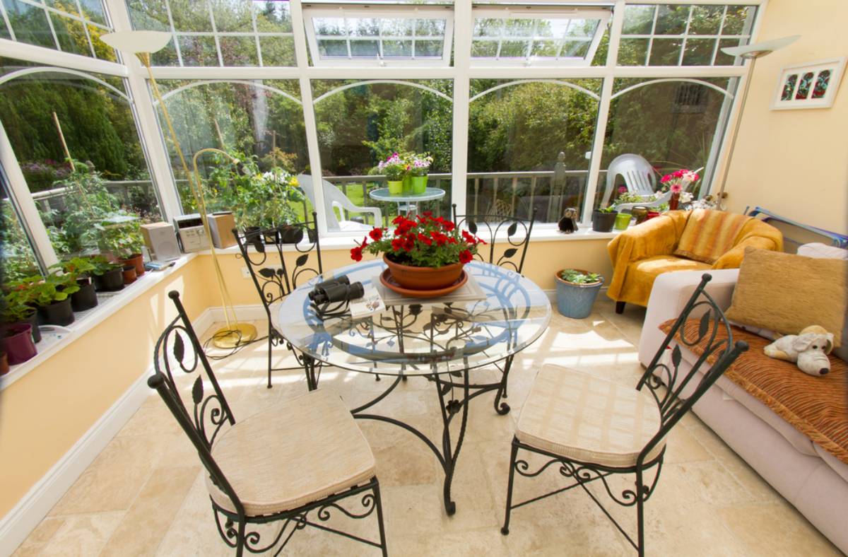 A bright all season sunroom with large windows, a round glass table with four chairs, and various potted plants. A cozy sofa with cushions and a stuffed toy sits in the corner as sunlight fills this inviting space.