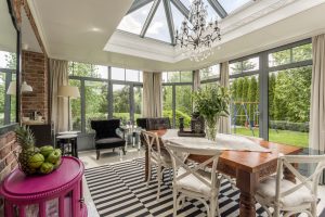 A sunlit dining room with large windows, a glass ceiling, chandelier, striped rug, and wooden table with white chairs sits in a Nassau home, featuring a purple side table and garden views with a swing set outside.