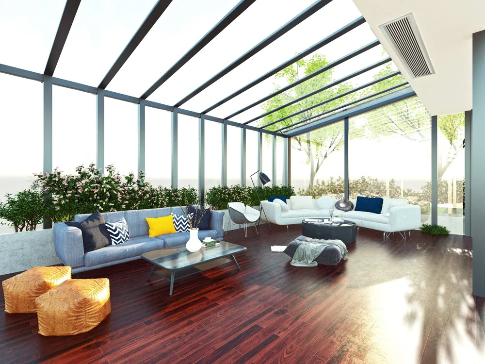 A bright, modern sunroom with floor-to-ceiling windows and a glass roof—perfect inspiration for sunrooms Long Island, NY. White and blue sofas, colorful cushions, plants, poufs, and dark wooden floors create a welcoming space filled with sunlight.