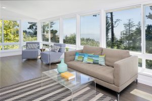 A modern living room by Sunroom Company Long Island with large windows, a gray sofa with colorful pillows, two armchairs, a clear coffee table with a vase, and a striped rug. Trees and distant water views enhance this inviting NY space.